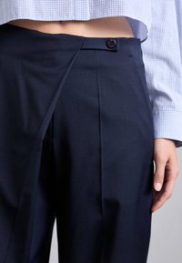 Marineblauwe broek met een gestructureerd ontwerp, voorzien van een vlakke voorkant, knoopsluiting en scherpe plooien. Glad materiaal met een getailleerde pasvorm.