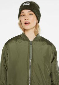 Grüne Bomberjacke mit Reißverschluss, geripptem Kragen und Seitentaschen. Kombiniert mit einer dunkelgrünen gestrickten Mütze mit Etikett.