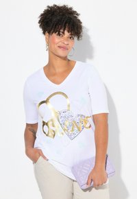 Camiseta blanca de cuello en V con gráficos de corazones dorados y brillantes. El modelo sostiene un bolso acolchado de color lavanda. Tez clara, cabello rizado y corto.