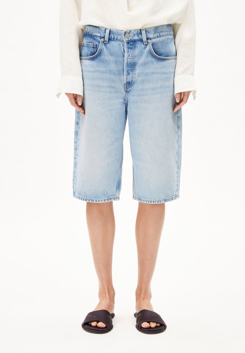 Hellblaue Jeansshorts mit lockerer Passform, fünf Taschen und einem Saum, der knapp über dem Knie endet. Kombiniert mit schwarzen Slippers.