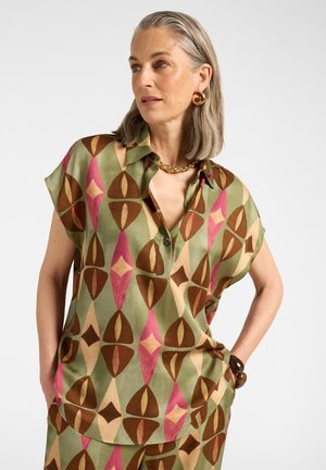 Femme aux cheveux gris portant un chemisier à manches courtes à motif géométrique vert, rose et marron ainsi qu'un pantalon assorti, collier en or et boucles d'oreilles.