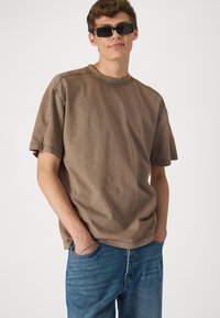 T-shirt en coton marron avec un col rond, des manches courtes et des détails de couture visibles, associé à un jean bleu et un bracelet en argent.