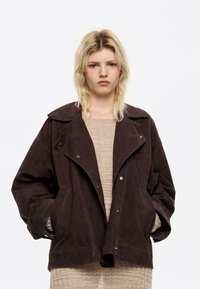 Bruine suède jas met brede revers, drukknoopsluiting en grote zakken. Boxy pasvorm met een textuurstof, gedragen over een beige gebreid top.