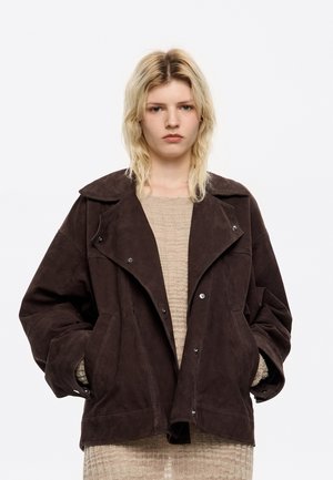 Bruine suède jas met brede revers, drukknoopsluiting en grote zakken. Boxy pasvorm met een textuurstof, gedragen over een beige gebreid top.