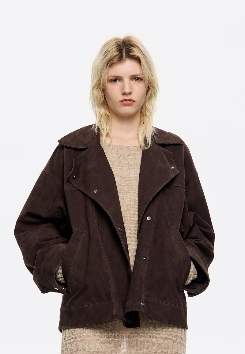 Bruine suède jas met brede revers, drukknoopsluiting en grote zakken. Boxy pasvorm met een textuurstof, gedragen over een beige gebreid top.
