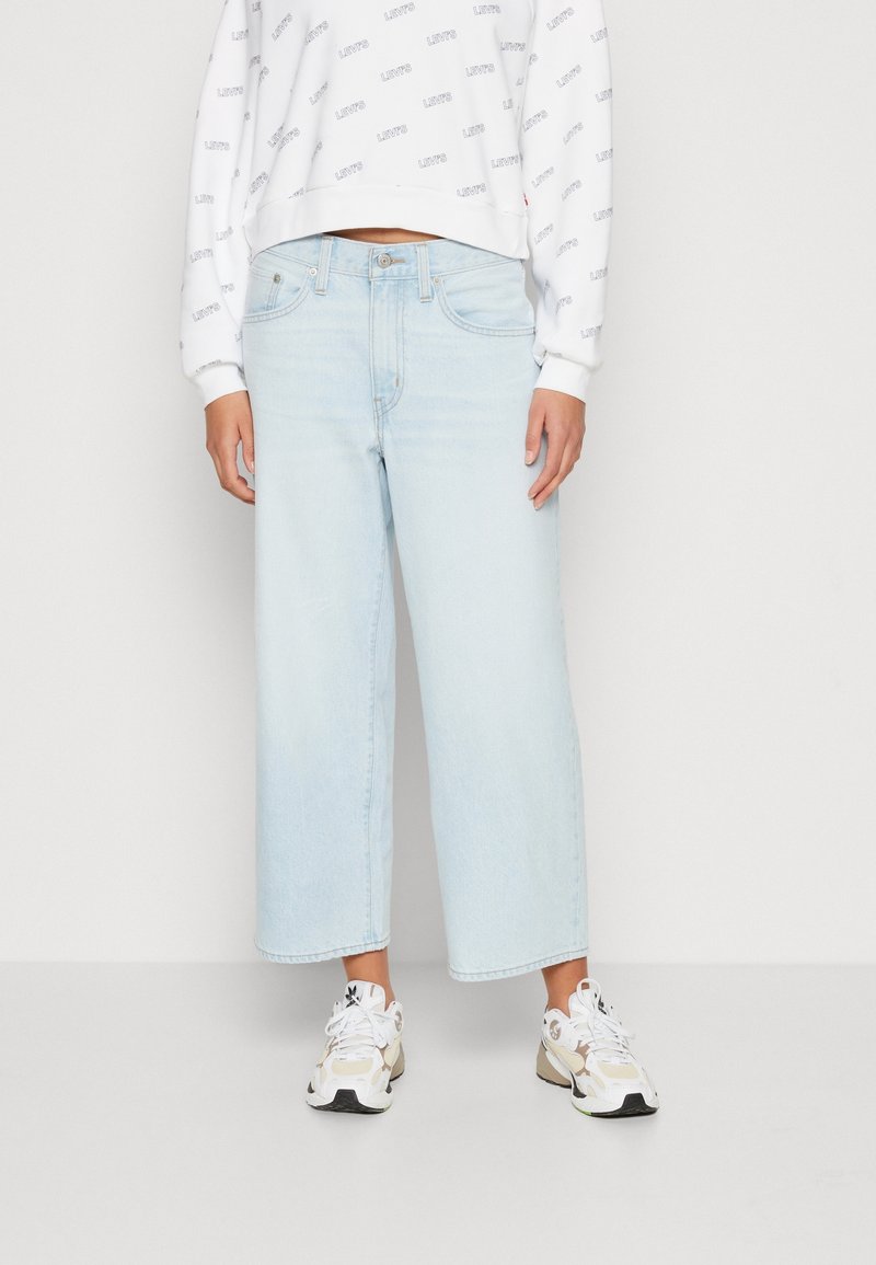 Personne vêtue d’un jean large court bleu clair, d’une chemise blanche à manches longues à motifs et de baskets blanches, debout sur un fond uni.