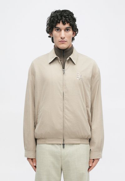 Ανοιχτό μπεζ φερμουάρ μπουφάν με γιακά, ελαστικούς μανσέτες και κεντημένο λογότυπο. Φοριέται πάνω από ένα καφέ ριγέ ζιβάγκο. Υποαλλεργικό ύφασμα.