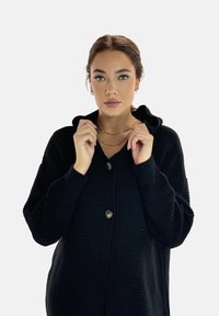 Svart stickad cardigan med huva, med ett texturerat mönster, två stora knappar och ribbad manchetter. Modellen håller i kragen.