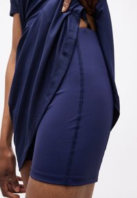 Persoon die een marineblauw shirt optilt om marineblauwe fietsbroekjes met zichtbare zijnaadsteken te tonen tegen een lichte achtergrond.