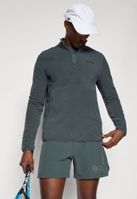 Pullover grigio in pile a maniche lunghe con zip a un quarto, caratterizzato da un pannello più chiaro a contrasto e dal logo 'BORG'. Abbinato a pantaloni corti grigi.