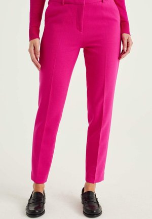 Felroze op maat gemaakte broek gedragen met zwarte loafers en een bijpassende felroze top met lange mouwen, zichtbaar van taille tot voeten.