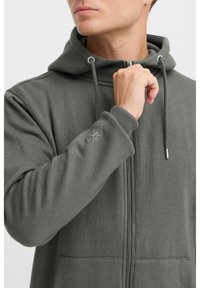 Olivgrüner Reißverschluss-Hoodie aus weichem Stoff, mit Kängurutasche, verstellbarem Kapuzenzug und gesticktem Detail am Ärmel.