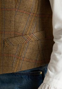 Hnedá károvaná vesta so štruktúrovanou tkaninou s modrými a červenými pruhmi, dvoma prednými vreckami a jemne prispôsobeným dizajnom. Nosená cez bielu blúzku.