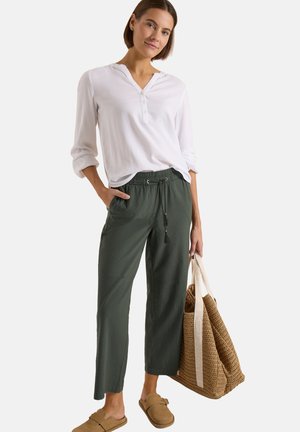 Femme portant un chemisier blanc boutonné et un pantalon vert foncé, tenant un grand sac tissé, debout avec une main dans la poche.