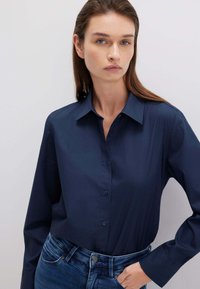 Camicia blu navy con bottoni, maniche lunghe, colletto classico, tessuto liscio, abbinata a jeans di denim blu. Focus sulle linee pulite e sul fit della camicia.