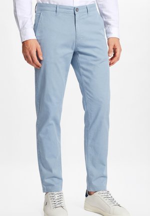 Mężczyzna ubrany w jasnoniebieskie proste spodnie, białą koszulę na guziki i białe sneakersy, stojący na białym tle.