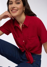 Blusa rossa a maniche corte con tessuto testurizzato, due tasche frontali e design con maniche legate alla vita, abbinata a jeans blu a vita alta.