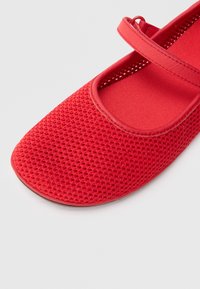 Zapato plano de malla roja con puntera redondeada, que presenta una correa ancha sobre el empeine y una parte superior de tela texturizada.