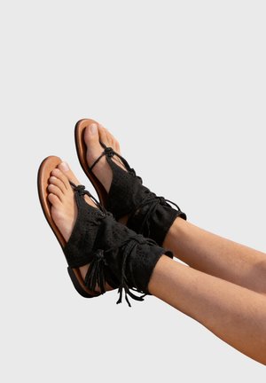 Jambes étendues portant des sandales en daim noir à lacets avec des détails perforés et des semelles couleur tabac sur un fond clair uni.