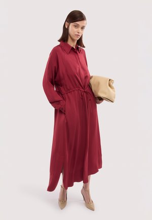 Kobieta w sukience na długi rękaw w kolorze czerwonym z paskiem ściągającym w talii, trzymająca beżową kopertówkę, nosząca beżowe buty na wysokim obcasie, na jednolitym jasnym tle.