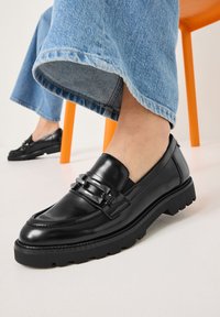 Zwarte leren instappers met een dikke zool, voorzien van dubbele gespen en textuuraccenten. Draag ze met lichtblauwe wijde jeans.