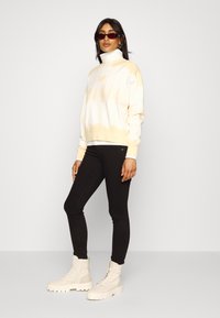 Sweatshirt em tie-dye branco e amarelo com gola alta, calças skinny pretas e botas de atacadores cremosas. Os acessórios incluem óculos de sol escuros.