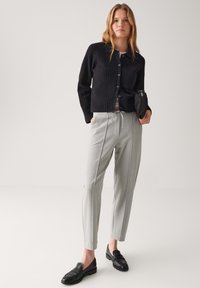 Jonge vrouw in een zwarte geribbelde cardigan, grijze tailored broek, zwarte loafers, met een opgevouwen zwarte paraplu tegen een effen achtergrond.