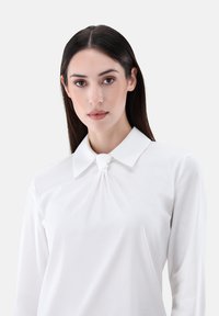 Weiße Bluse mit klassischem Kragen und einem geknoteten Detail vorne, mit glatter Textur und langen Ärmeln.