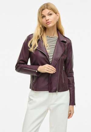 Giacca in pelle borgogna con zip frontale asimmetrica, colletto a revers e tasche con zip, abbinata a una maglietta a righe e pantaloni bianchi.