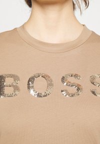Beżowy t-shirt z metalicznymi cekinami w kształcie napisu "BOSS", okrągłym dekoltem, krótkimi rękawami i miękkim połączeniem bawełny.