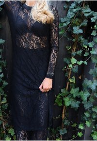 Fekete csipkeruha hosszú ujjakkal, virágmintákkal és áttetsző felső réteggel. Az anyag finom textúrájú, aláöltöző szoknyával megtervezve.
