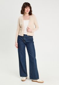 Cardigan en maille beige avec un col en V, associé à un jean en denim foncé à jambes larges et des baskets claires. Design simple, texture lisse.