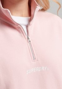 Sudadera de forro polar de color rosa claro con cremallera, cuello alto, con cremallera plateada y el logo blanco "SUPERDRY" en el pecho. Textura suave.