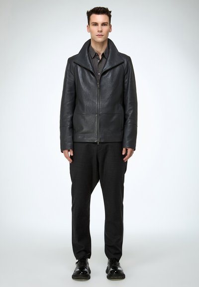 Schwarze Lederjacke mit Reverskragen und zwei Fronttaschen, getragen über einem dunklen Hemd und kombiniert mit locker sitzenden Hosen und schwarzen Schuhen.