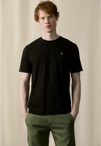Zwarte katoenen t-shirt met een ronde hals en korte mouwen, voorzien van een klein wit logo aan de linkerkant. Gekocht met groene broek.