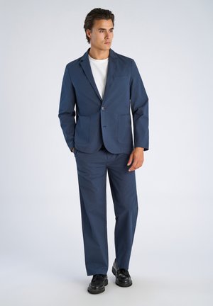 Man in een donkerblauw pak met een colbert met twee knopen en wijde broekspijpen, gecombineerd met een wit T-shirt en zwarte schoenen, staand tegen een grijze achtergrond.