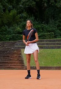 Joueuse de tennis portant un t-shirt noir à manches courtes et une jupe plissée blanche, tenant une raquette. Chaussettes et chaussures noires, arrière-plan d'un court en terre battue en extérieur.