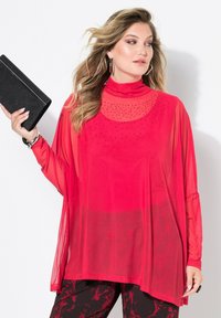 Rotes halbtransparentes Oberteil mit langen Ärmeln, hohem Kragen und verzierten Details. Hält eine schwarze Clutch mit glitzernder Textur.