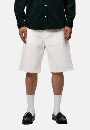 Persona che indossa una camicia nera con bottoni, pantaloncini bianchi larghi fino al ginocchio, calzini bianchi e mocassini neri, in piedi davanti a uno sfondo semplice.