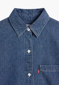 Chemise en denim bleu à boutons avec col, boutons blancs, poche poitrine et étiquette de marque rouge. Le tissu a une texture classique.