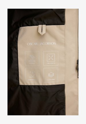 Väriä tan ja musta sisältävä takki, jossa on tasku, jossa on valkoisia kuvastoja, jotka ilmoittavat tuulenpitävyydestä, vedenpitävyydestä, neljän suuntaisen venymisestä ja kolmitasoisesta kankaasta.