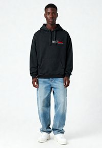 Felpa nera con cappuccio con la scritta "SWEET" in rosso e bianco, tasca a marsupio, polsini a coste, abbinata a jeans blu chiaro a vestibilità rilassata e sneakers bianche.