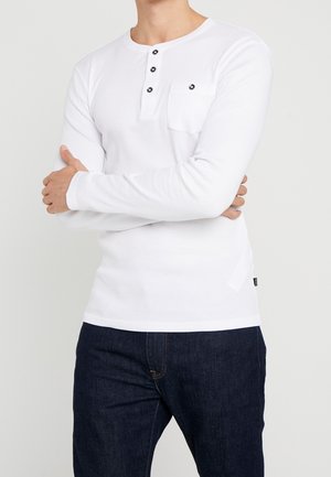 Homme portant une chemise Henley blanche à manches longues avec des boutons noirs et un jean bleu foncé, bras croisés sur la poitrine.