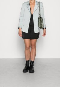 Vaaleanharmaa kaksirivinen bleiseri, musta nappikauluspaita ja mustat kumiset nilkkasaappaat. Malli pitää kädessään vihreää tilkkulaukkua.