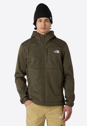 Olive green softshell jacket with a zippered front, adjustable hood, and logo on the chest; paired with tan pants. Black beanie worn.