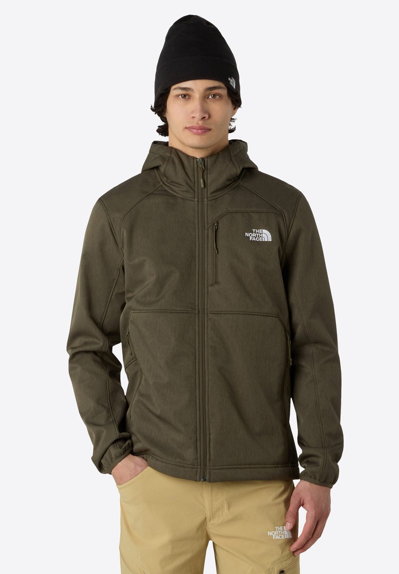 Oliwkowa kurtka softshell z zamkiem z przodu, regulowanym kapturem i logo na piersi; połączona z beżowymi spodniami. Na głowie czarna czapka.