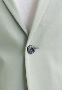 Lys grønn blazer med teksturert overflate, med en enkelt svart knapp og notched revers. Hvit innvendig fôr synlig.