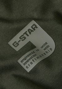 Olijfgroen stof met een gestructureerd, rechthoekig label met reliëftekst, waaronder "G-STAR" en "ontwikkeld om te verenigen met vakmanschap."