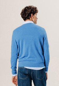Blauer Kaschmirpullover mit Rundhalsausschnitt, langen Ärmeln und geripptem Saum, getragen über einem hellblauen gestreiften Hemd, kombiniert mit dunklen Jeans.