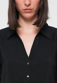 Blusa preta com decote em V, textura subtil e botões dourados à frente. Ajuste sob medida com colarinho clássico.